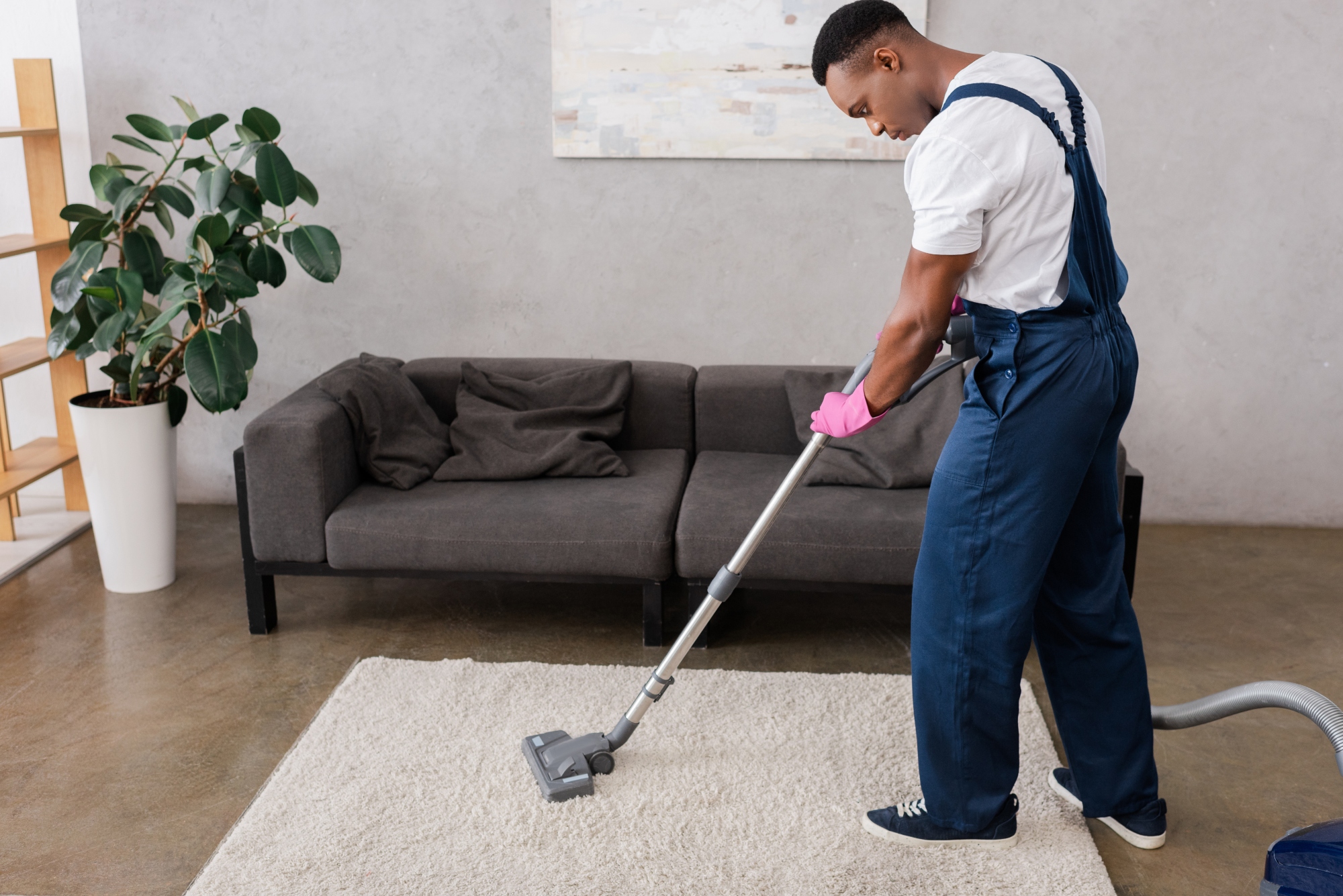 African american cleaner in overalls and rubber glove cleaning carpet while working in living room